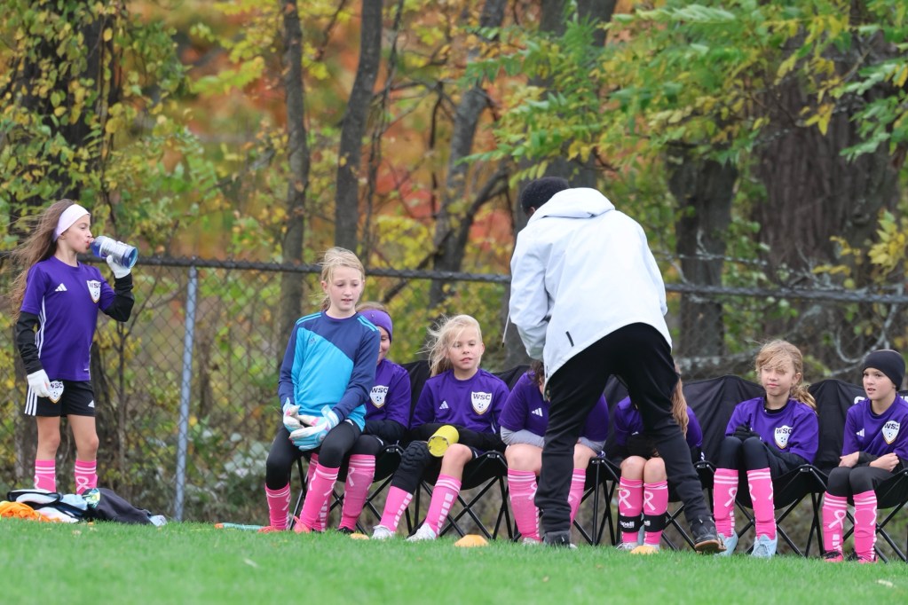 A group of young female soccer players in purple jerseys sitting on benches during a team meeting, with one player drinking water and a coach addressing them.