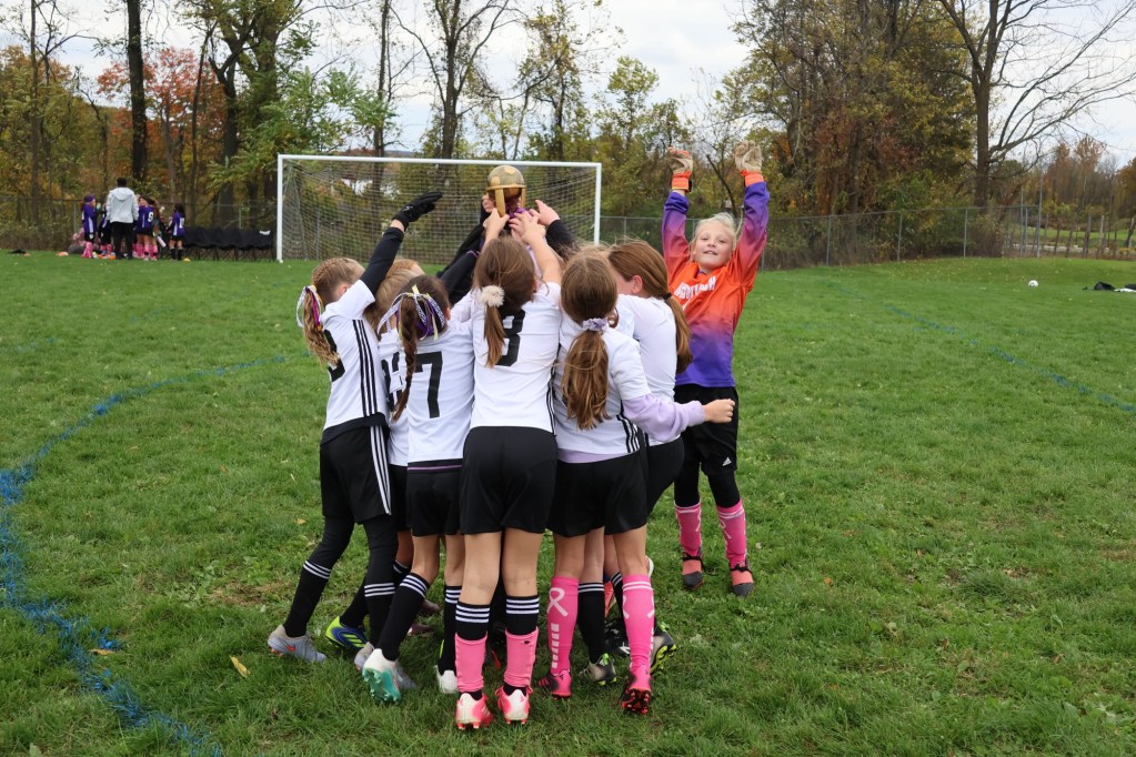 A group of young girls in soccer uniforms celebrating and lifting a trophy on a grassy field.