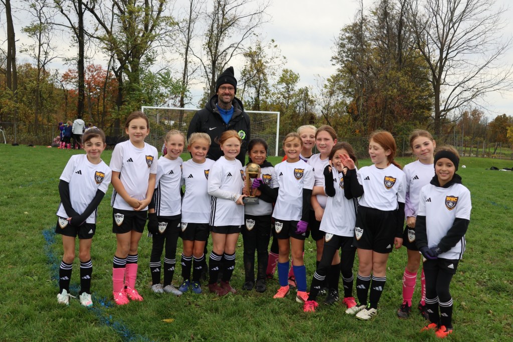 A youth soccer team poses for a photo with their coach on a grassy field, holding a trophy. The team is wearing white jerseys with black shorts and black socks, and they appear to be celebrating.
