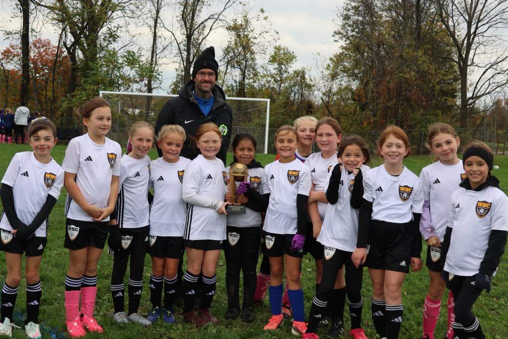 A group of young girls wearing soccer uniforms, standing together on a grassy field, with an adult coach holding a trophy. The girls are smiling and celebrating their victory, with trees in the background.