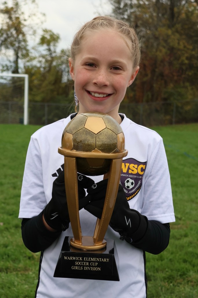A smiling girl in a soccer uniform holding a trophy with a soccer ball on top, commemorating the Warwick Elementary Soccer Cup, Girls Division.