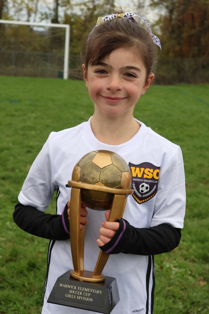 A young girl in a soccer uniform holds a trophy with a soccer ball on top, smiling proudly. The trophy is labeled 'Warwick Elementary Soccer Cup Girls Division.'