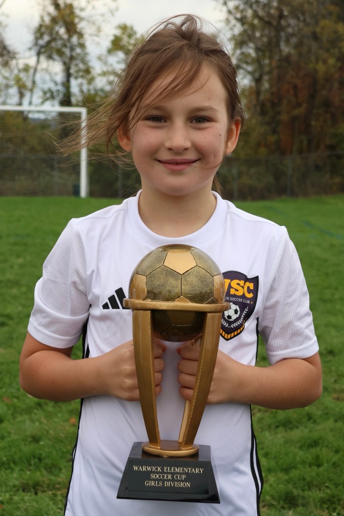A young girl in a white soccer jersey holding a trophy with a soccer ball on top, smiling outdoors on a soccer field.