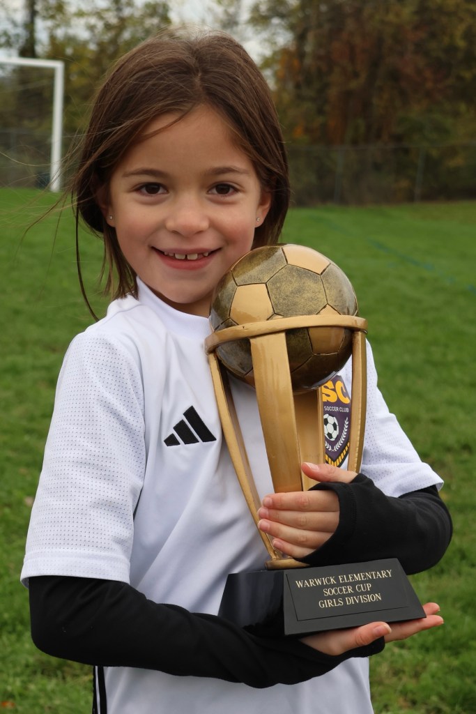 A young girl in a soccer uniform holding a trophy with a soccer ball on top, smiling proudly on a grass field.