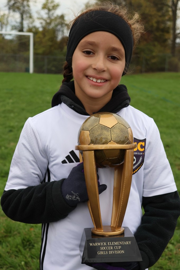 A young girl wearing a soccer jersey and gloves holds a trophy with a golden soccer ball on top, celebrating her win at the Warwick Elementary Soccer Cup in the Girls Division.