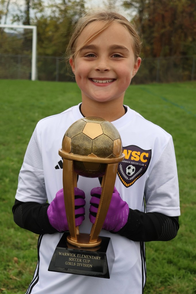 A young girl in a soccer uniform proudly holds a trophy with a soccer ball on top, celebrating her achievement in the Warwick Elementary Soccer Cup Girls Division.