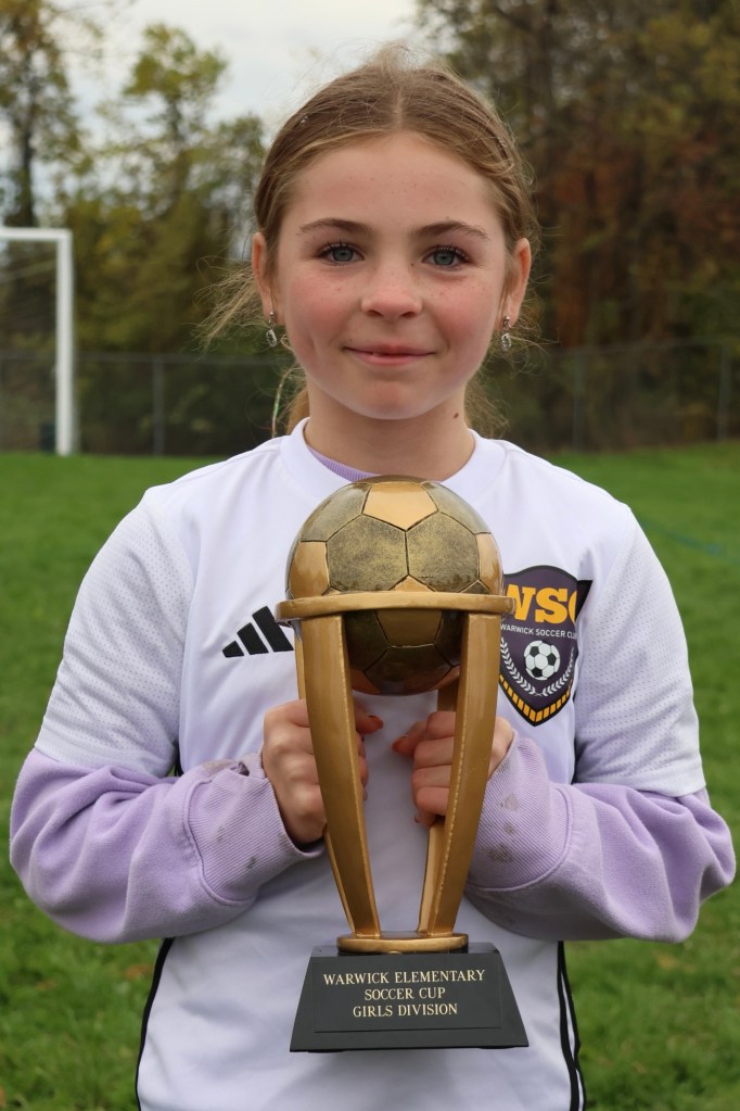 A young girl in a soccer uniform holding a trophy with a soccer ball on top, standing on a green field.