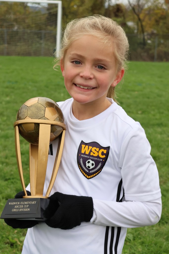 A young girl in a soccer uniform proudly holding a trophy with a soccer ball on top, standing on a grassy field with a soccer goal in the background.