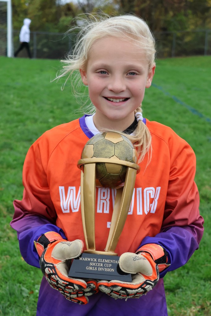 A young girl wearing an orange and purple soccer jersey and gloves smiles while holding a trophy that reads 'Warwick Elementary Soccer Cup Girls Division.'